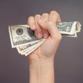 Woman hand squeezes dollar bills against grey background.