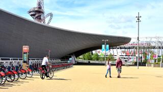 Person riding from Santander bike hire docking station at Aquatic Centre with Orbit Tower beyond in Queen Elizabeth Olympic Park Stratford London UK