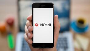 A man looks at his iPhone which displays the UniCredit logo, while sat at his computer desk (Editorial use only).