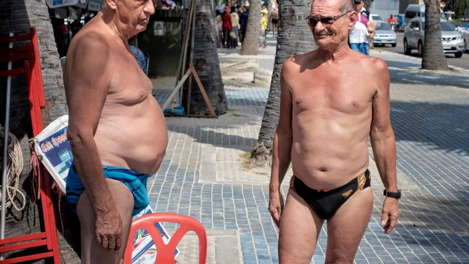 Two mature men with pronounced stomachs and wearing speedos swimming trunks chatting on the street