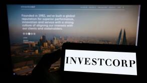 Person holding mobile phone with logo of Bahraini company Investcorp Holdings B.S.C. on screen in front of web page. Focus on phone display.