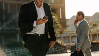 Businessman looking at his mobile phone while walking on street to office. Busy office going people carrying office bag and using mobile phone.