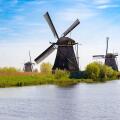 Panoramic view of multiple historic Dutch windmills lining the waterways in Kinderdijk, Netherlands, surrounded by green fields under a vast blue sky.