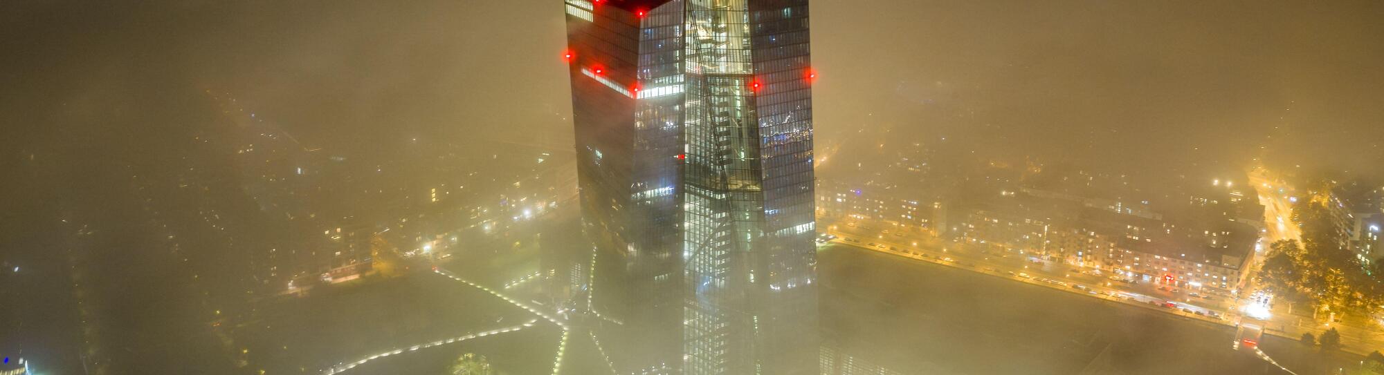 Aerial view of the European Central Bank skyscraper piercing through a sea of fog, its red lights ablaze against the muted city lights, Frankfurt am M
