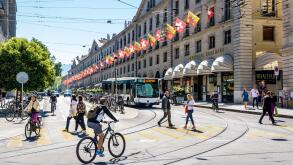 Pedestrians and cyclists are crossing the rue de la Corraterie in Geneva, with buildings decked with flags, as a bus is waiting at a red light.