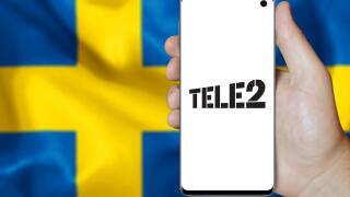 A man holds a smartphone displaying the logo of company Tele2 listed on OMX Stockholm. Swedish flag in the background. Credit: PIXDUCE