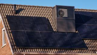 A modern, energy-efficient house in Assen, Netherlands, featuring integrated heat pumps on the roof, solar panels, and a clear blue sky.