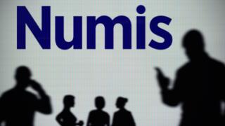 The Numis Securities logo is seen on an LED screen in the background while a silhouetted person uses a smartphone (Editorial use only)