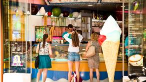 three young women queuing at ice cream parlor purchasing ice cream cones, girl in shorts holding her head as if she was struggling to make a choice.