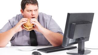 Stop showing me healthy things. A young man eating his lunch at his desk at work while staring with mouth agape at his monitor - unhealthy eating habits.