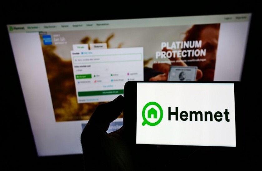 Person holding smartphone with logo of Swedish property platform company Hemnet AB on screen in front of website. Focus on phone display.