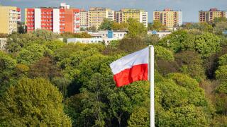 Polish state flag in the background of sunlit green trees in autumn. In the distance is a row of colorful new high-rise houses and a blue sky with clo