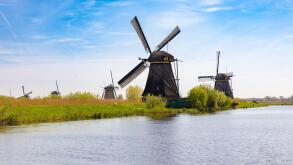 Panoramic view of multiple historic Dutch windmills lining the waterways in Kinderdijk, Netherlands, surrounded by green fields under a vast blue sky.