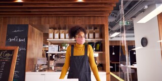 Female Small Business Owner Of Coffee Shop In Mask Standing Behind Counter During Health Pandemic