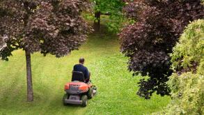 High angle view of a man using a ride-on mower to cut the grass in his back yard viewed between leafy green trees in spring or summer