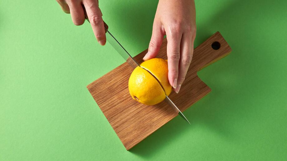 A woman's hands cut a yellow ripe lemon in half with knife on a wooden board on a green background. Top view.