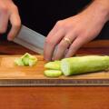 A male hand slicing a cucumber on a wooden cutting board.