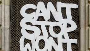 Graffiti with the message 'Can't Stop Won't Stop' on a wire mesh window grille in a historic stone doorway in Barcelona, Spain.