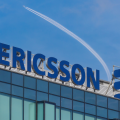 A plane flies overhead of a signboard featuring Ericsson's logo on the roof of one its corporate offices on the roof of a building on a sunny day. The Ericsson brand name or logo is set against a blue sky with a trail from a flying plane.