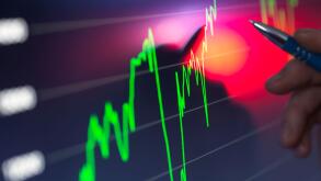 A hand holding a pen points to a bright stock market chart on a digital screen with fluctuating green and red graph lines indicating financial data.