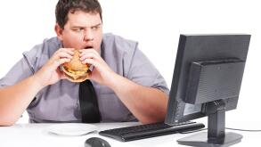 Stop showing me healthy things. A young man eating his lunch at his desk at work while staring with mouth agape at his monitor - unhealthy eating habits.