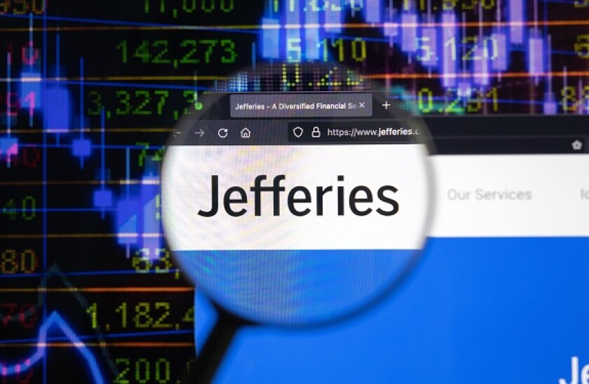 Jefferies company logo on a website with blurry stock market developments in the background, seen on a computer screen through a magnifying glass.