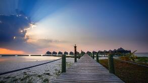 Sun bursting through clouds at dawn over water villas in the Maldives.