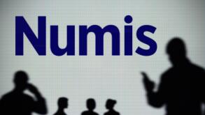 The Numis Securities logo is seen on an LED screen in the background while a silhouetted person uses a smartphone (Editorial use only)