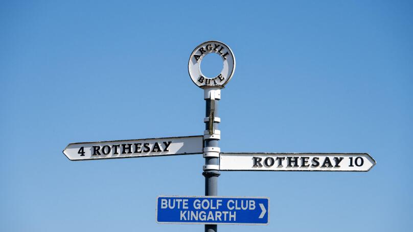 An island road sign on the Isle of Bute, Scotland against a blue sky. It shows the same destination, Rothesay, in two directions.