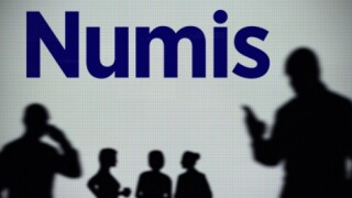 The Numis Securities logo is seen on an LED screen in the background while a silhouetted person uses a smartphone (Editorial use only)