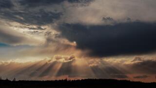 A breathtaking landscape showcasing dramatic crepuscular rays as the sun pierces through thick, moody clouds over a forested horizon.