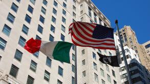 Flags of the United States, Italy, and POW/MIA waving in the wind against a backdrop of New York City buildings and a clear blue sky on a sunny day