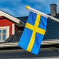 Close up photo of Swedish flag, blurry black roof and windows at top of Swedish wooden red house at background. Blue sky at sunny day