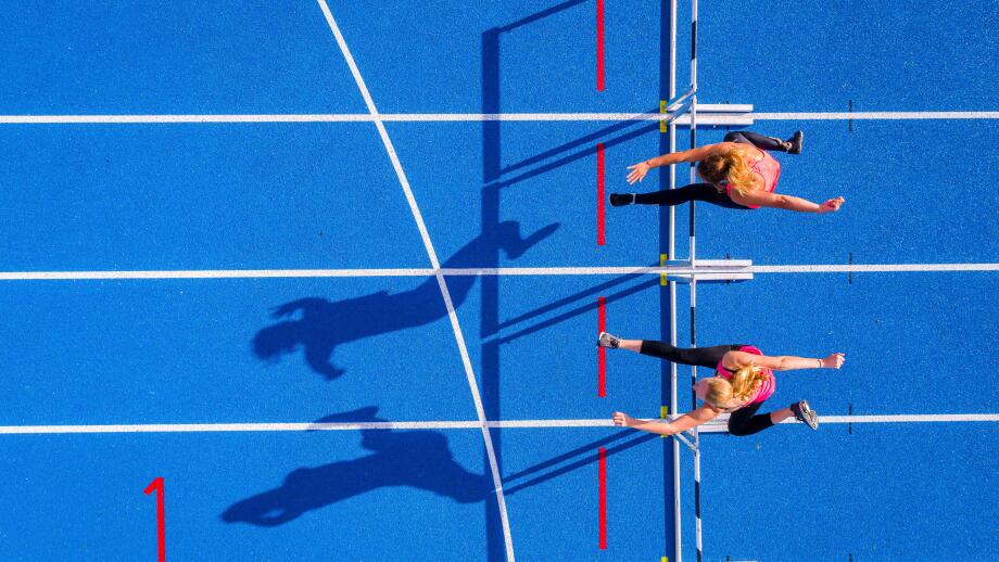 Top view of two female runners crossing hurdles on tartan track