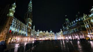 Night view of the illuminated Grand Place in Brussels, Belgium, with historic buildings and crowds gathered in the square.