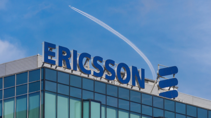 A plane flies overhead of a signboard featuring Ericsson's logo on the roof of one its corporate offices on the roof of a building on a sunny day. The Ericsson brand name or logo is set against a blue sky with a trail from a flying plane.