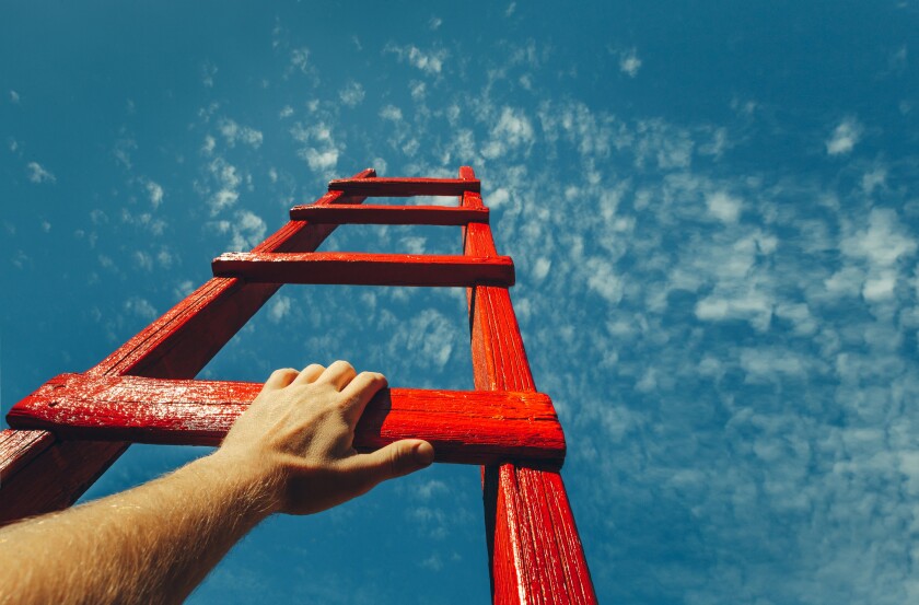 A male hand holds onto the crossbar of a red wooden staircase leading to the blue sky