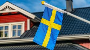 Close up photo of Swedish flag, blurry black roof and windows at top of Swedish wooden red house at background. Blue sky at sunny day
