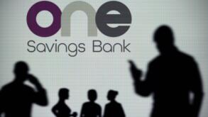 The OneSavings Bank logo is seen on an LED screen in the background while a silhouetted person uses a smartphone (Editorial use only)