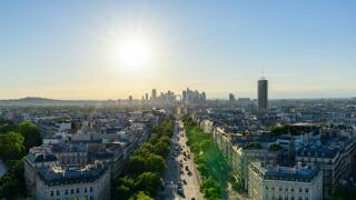 Sweeping aerial view along Avenue de la Grande Armee in Paris, framed by classic Haussmann buildings and green trees, with the modern towers of La Defense silhouetted against a golden evening sky.