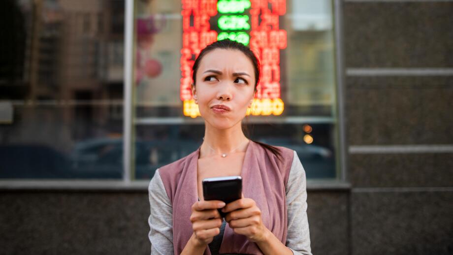 A young beautiful asian woman using an application in her smart phone to check currency exchange rates in front of an illuminated information board.