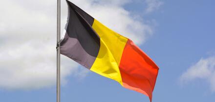 The national flag of Belgium flies up a flagpole on a beautiful summer day with a background of blue sky and white clouds.