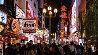 Crowd of tourists strolling down one of the streets in Osaka's Dotonbori area. Cityscape by night with lots of billboards and lights.
