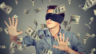 Blindfolded young businessman trying to catch dollar bills banknotes flying in the air isolated on gray wall background. Financial corporate success o