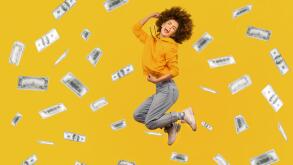 Portrait of successful rich woman with Afro hairstyle jumping in money rain, celebrating her victory, clenched fists and screaming happily. Indoor studio shot isolated on yellow background.