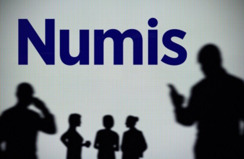 The Numis Securities logo is seen on an LED screen in the background while a silhouetted person uses a smartphone (Editorial use only)