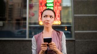 A young beautiful asian woman using an application in her smart phone to check currency exchange rates in front of an illuminated information board.