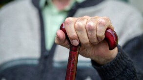 An elderly old age pensioner sits with his hands on a walking stick in a care home.