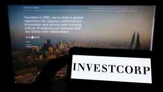 Person holding mobile phone with logo of Bahraini company Investcorp Holdings B.S.C. on screen in front of web page. Focus on phone display.