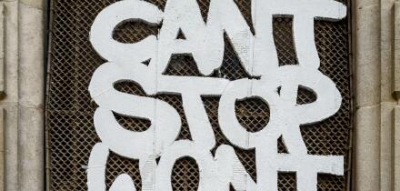 Graffiti with the message 'Can't Stop Won't Stop' on a wire mesh window grille in a historic stone doorway in Barcelona, Spain.